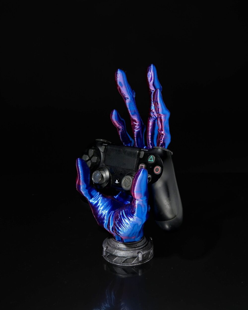 Alien hand-shaped stand in blue and pink hues holding a black gaming controller, displayed against a black background.