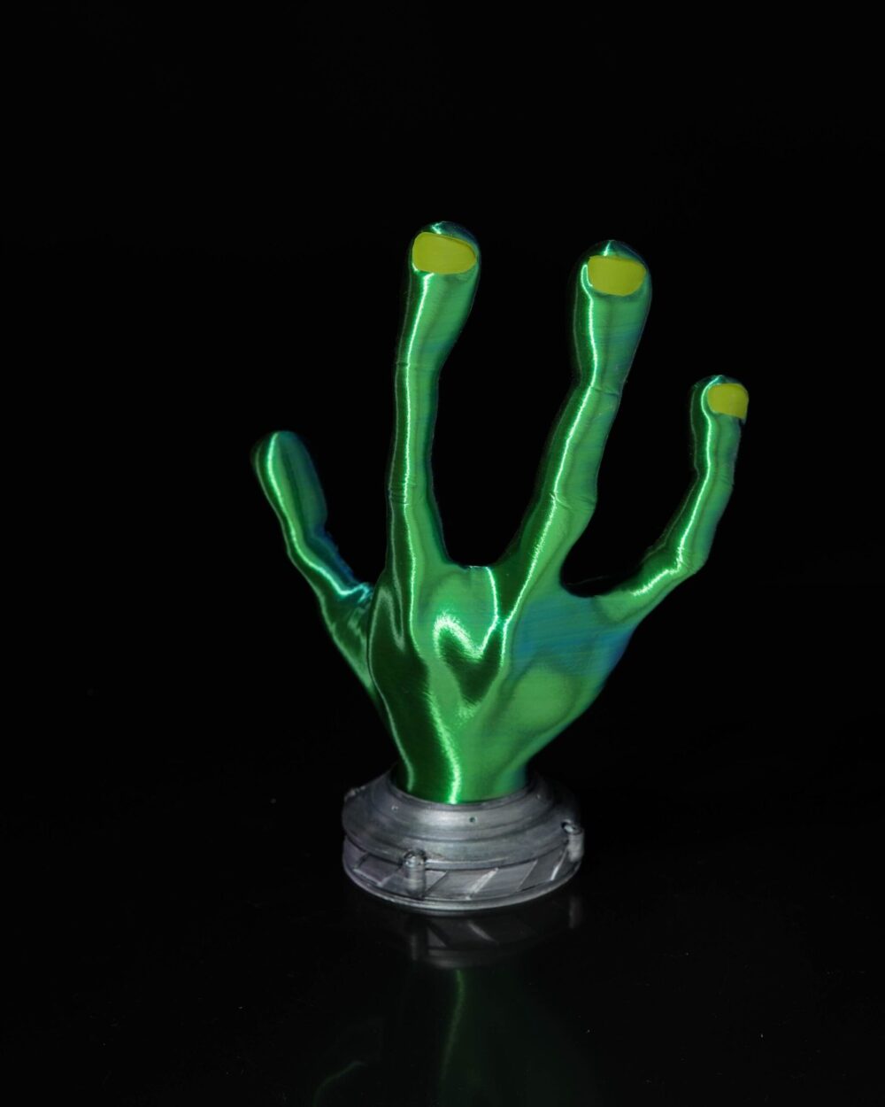 4-fingered green alien hand controller holder with yellow fingertips on a gray base, shown against a black background.