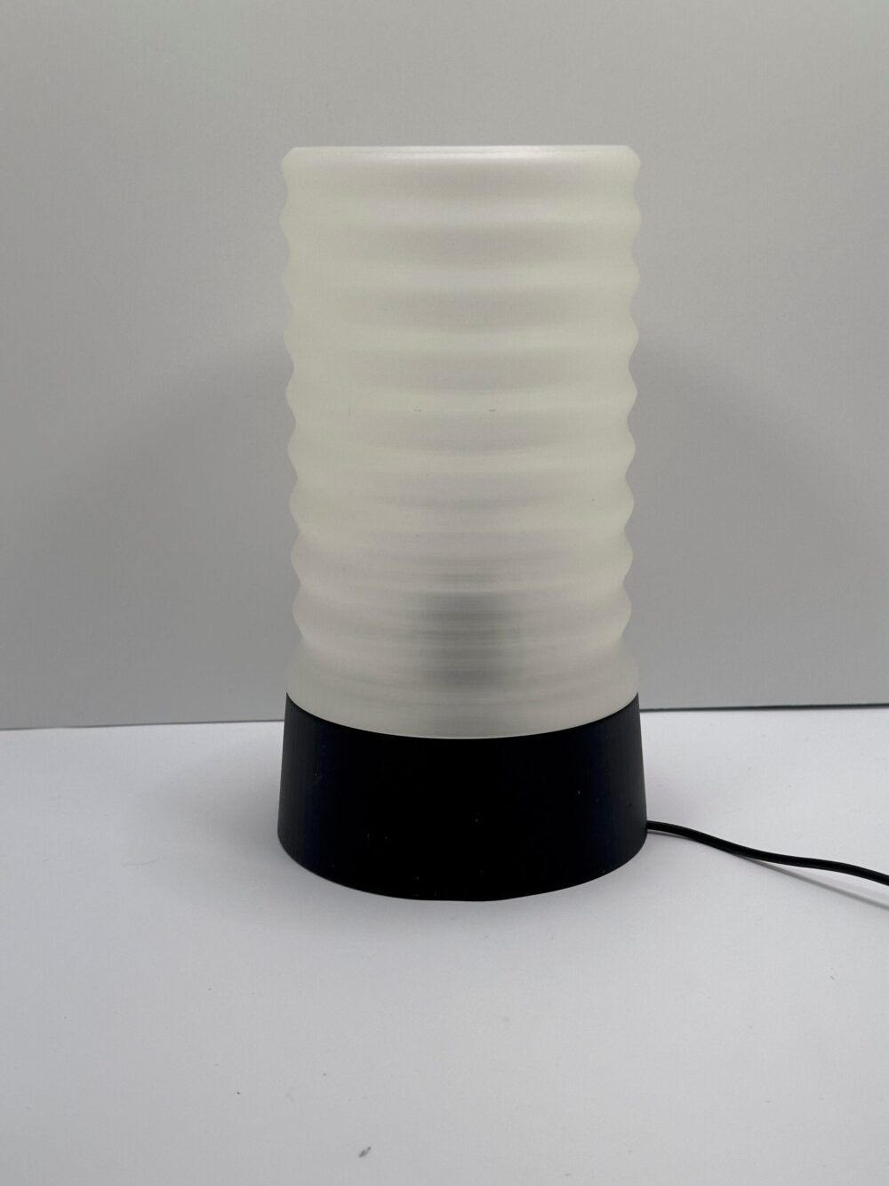 The Wavy Table Lamp features a frosted cylindrical shade with horizontal wave patterns, set on a sleek black base.