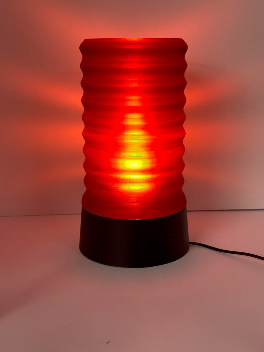 The Wavy Table Lamp emits a warm red glow, featuring a ribbed cylindrical design atop a sleek black base.